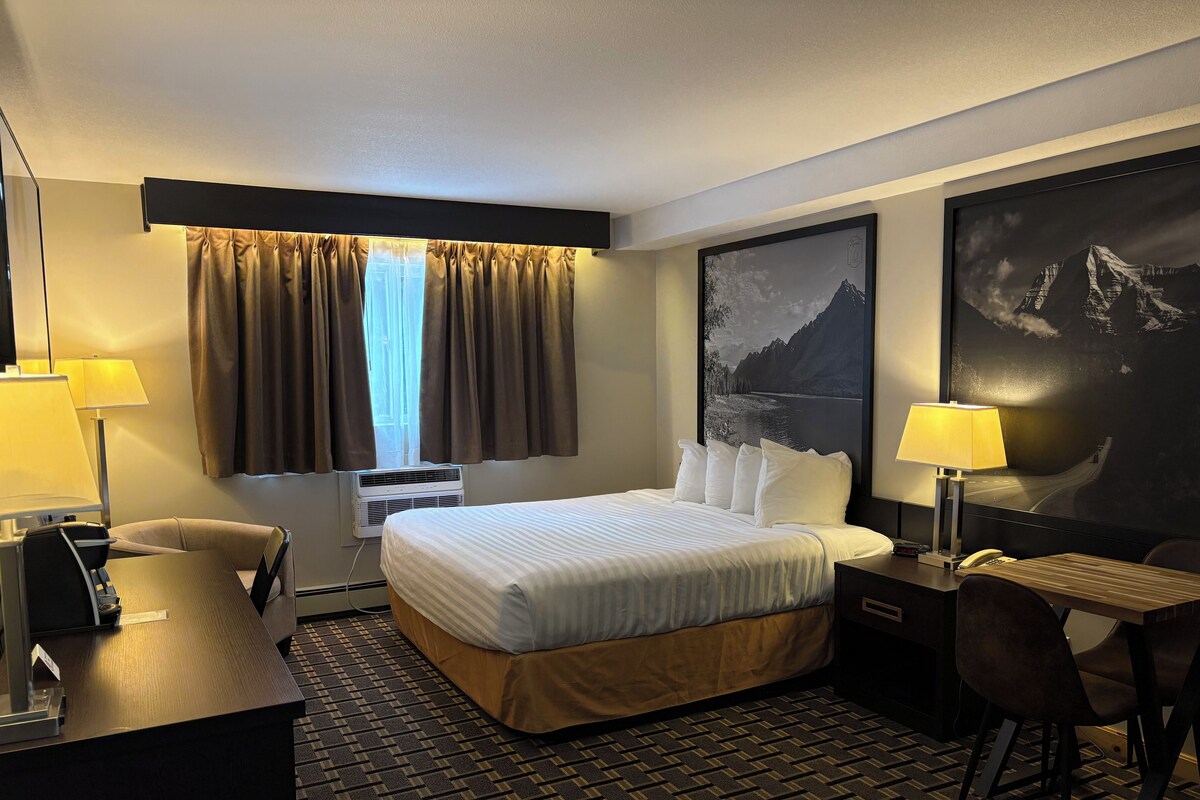Una habitación de hotel con una cama, un escritorio, una silla, una lámpara, una ventana con cortinas, aire acondicionado y un cuadro grande en la pared.