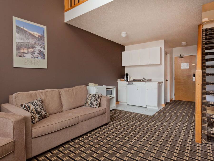 A living room with a sofa, a patterned rug, a kitchen area with white cabinets, and a bookshelf.