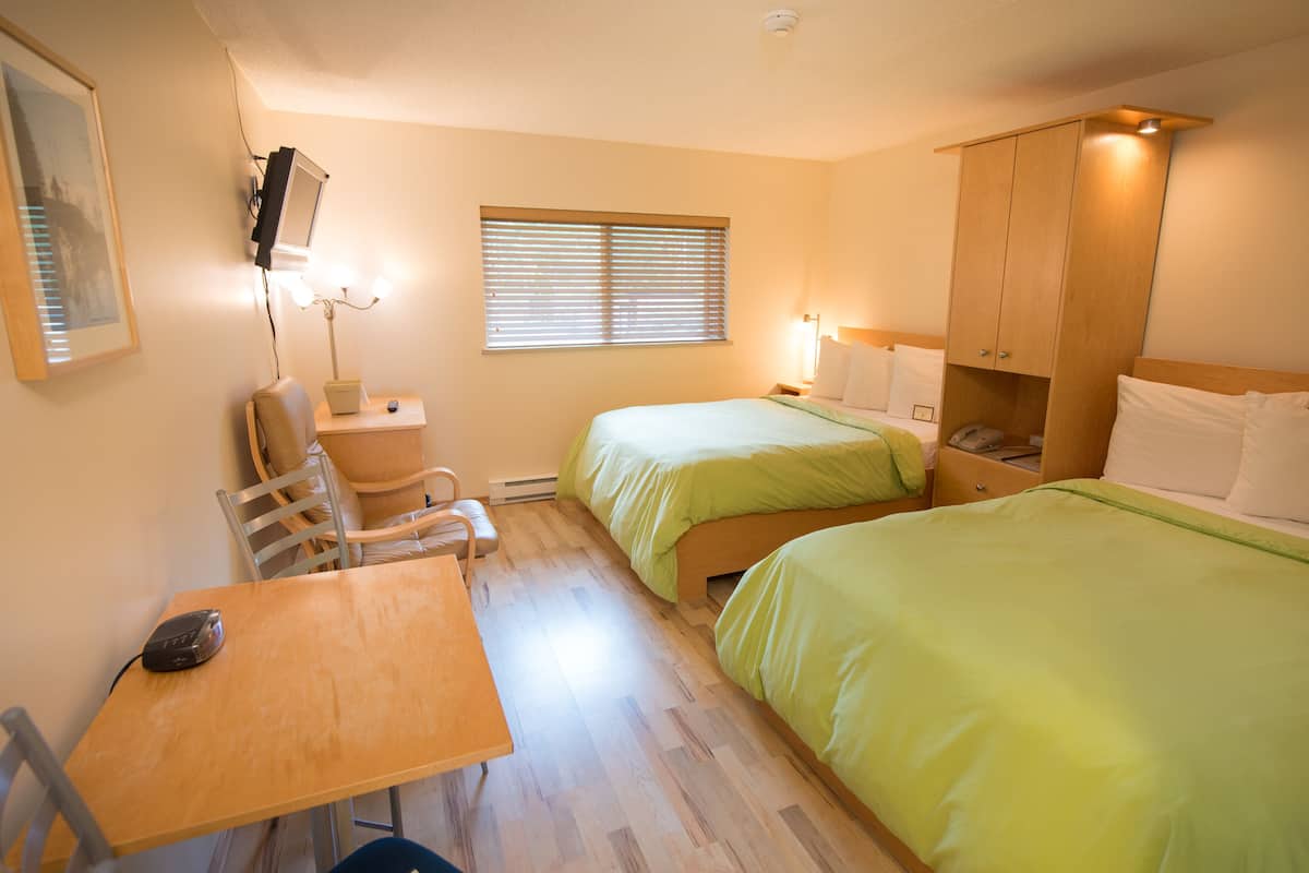 A hotel room with two beds, a desk, a chair, a small table, a lamp, and a window with blinds.