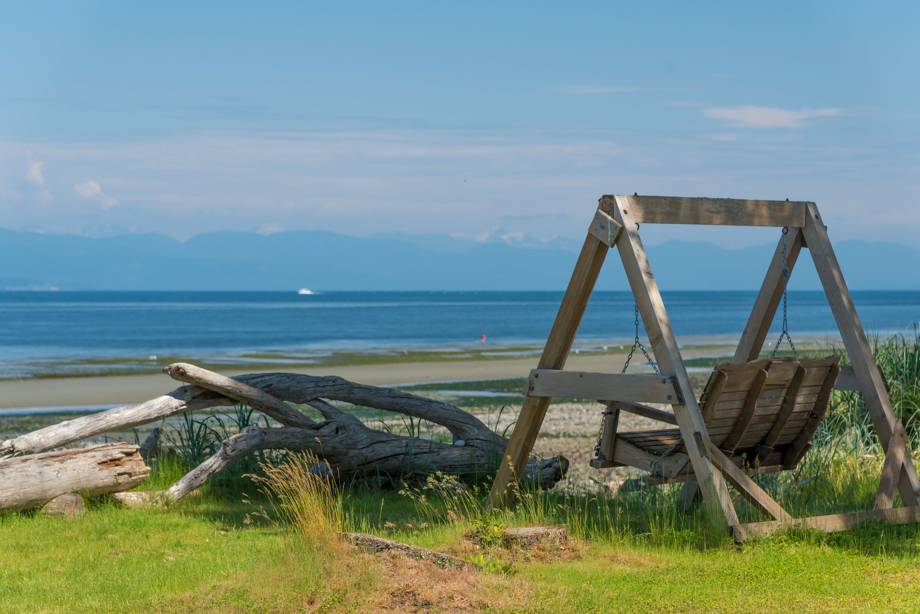 A wooden swing chair on a grassy area near a beach with a view of the ocean and mountains in the background.