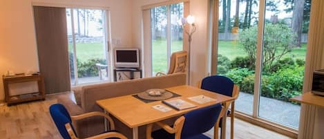 Una sala de estar moderna con una mesa de madera, sillas azules, un sofá, una televisión y ventanas amplias con vistas a un jardín.