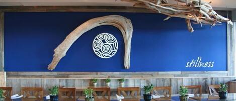 A restaurant interior with wooden tables, chairs, and a blue wall with a decorative piece and the word "stillness".