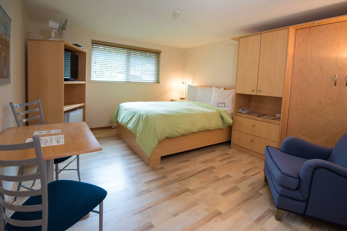 A compact bedroom with a bed, a desk, a chair, a wardrobe, and a window with blinds.