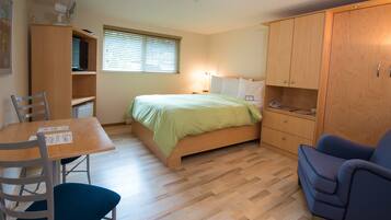 A compact bedroom with a bed, a desk, a chair, a wardrobe, and a window with blinds.