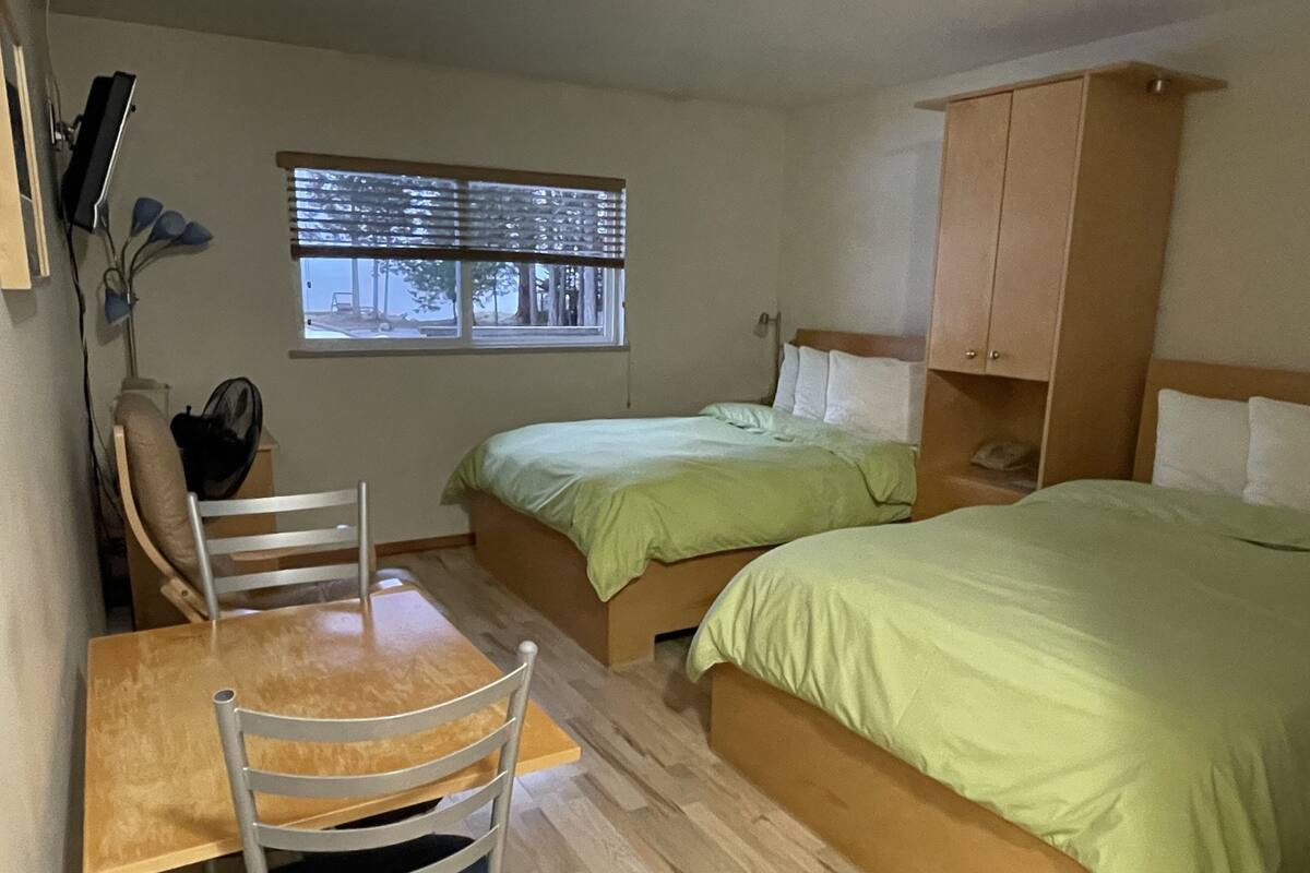 A room with two beds, a wooden table, a chair, a window with blinds, and a wooden wardrobe.