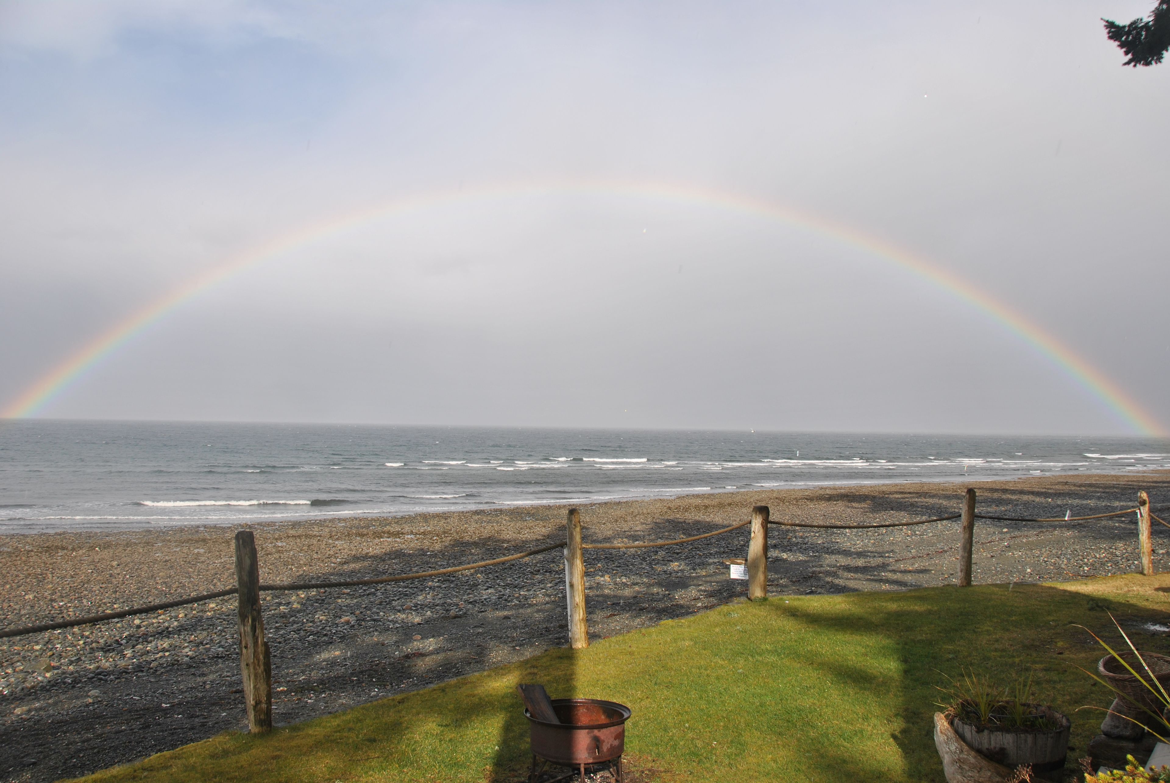 Une plage avec un arc-en-ciel, une clôture en bois et une zone herbeuse avec un pot de fleurs.