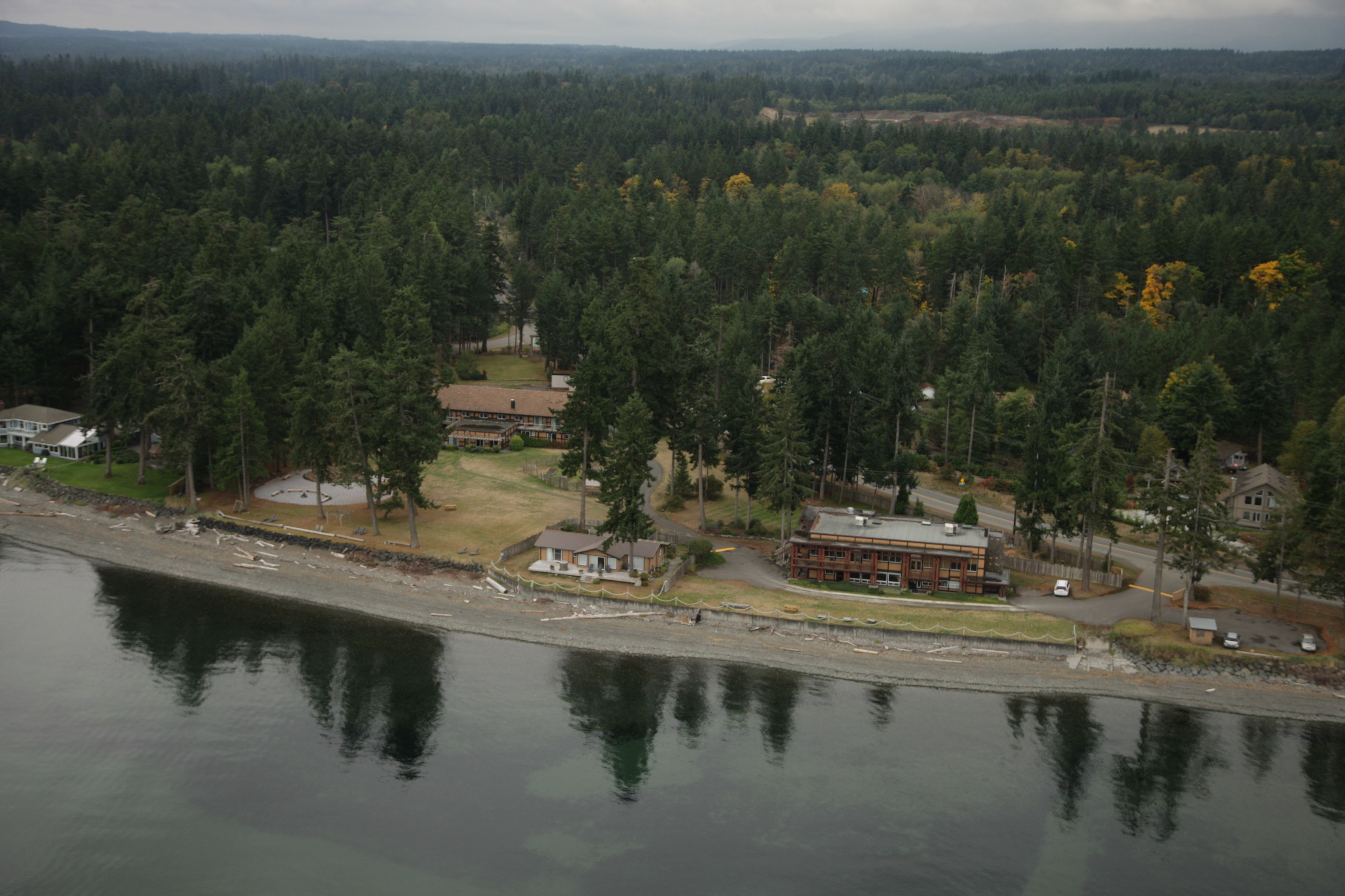 Vista aérea de una propiedad junto al lago con varias construcciones, incluyendo un gran edificio principal y casas más pequeñas, rodeadas de un bosque denso.