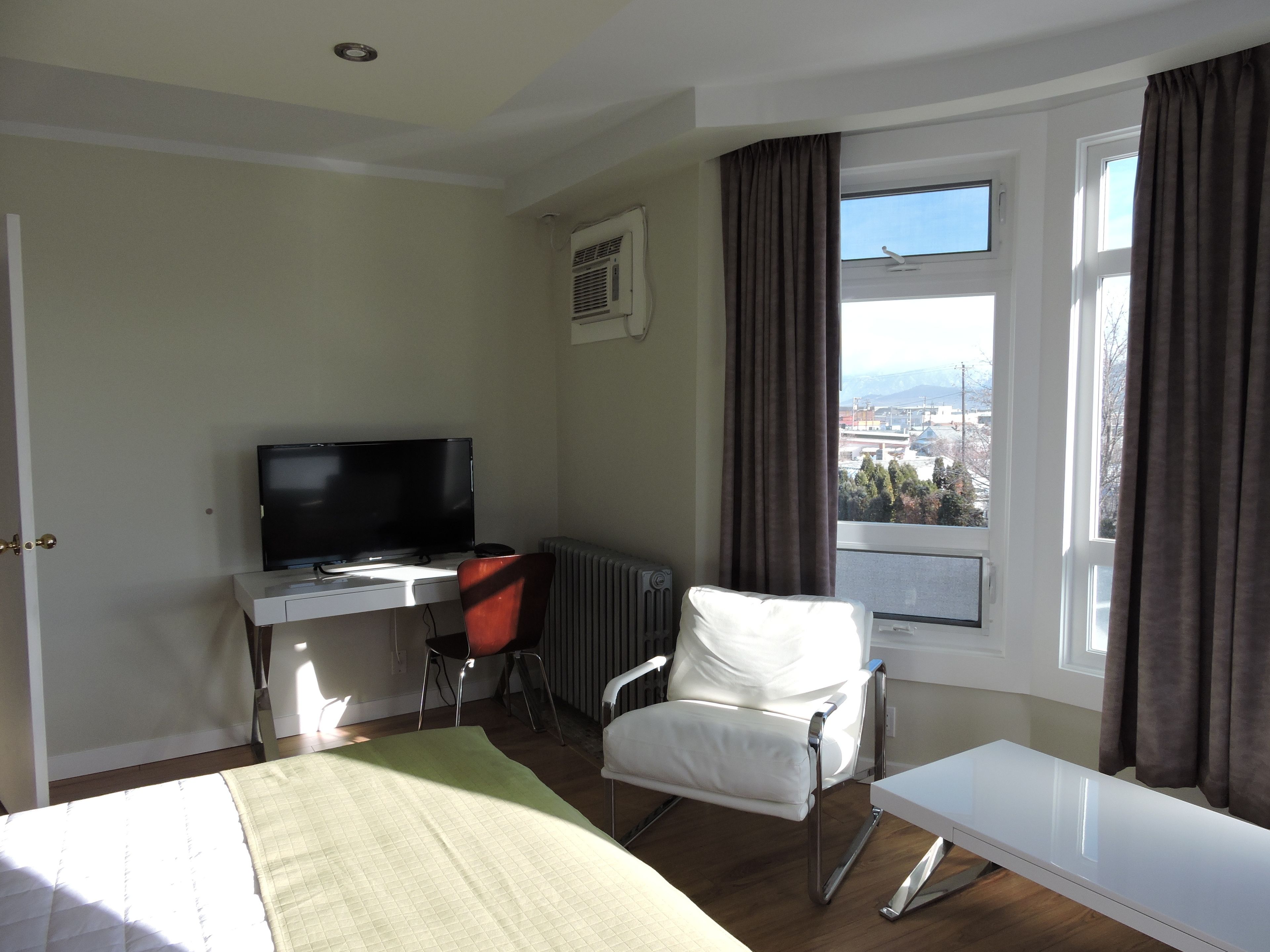 Habitación de hotel con cama, escritorio con computadora, silla, ventana con cortinas y radiador.