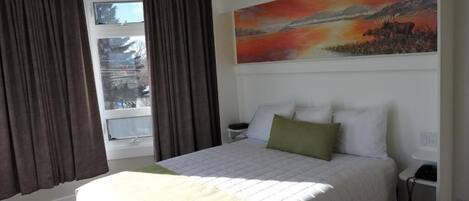 A hotel room with a large bed, a painting of a sunset, a window with curtains, and a bedside table.