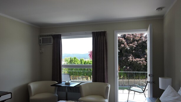 A hotel room with a bed, two chairs, a small table, and a view of a balcony with a railing and trees. - Almo Court Motel (Cranbrook)