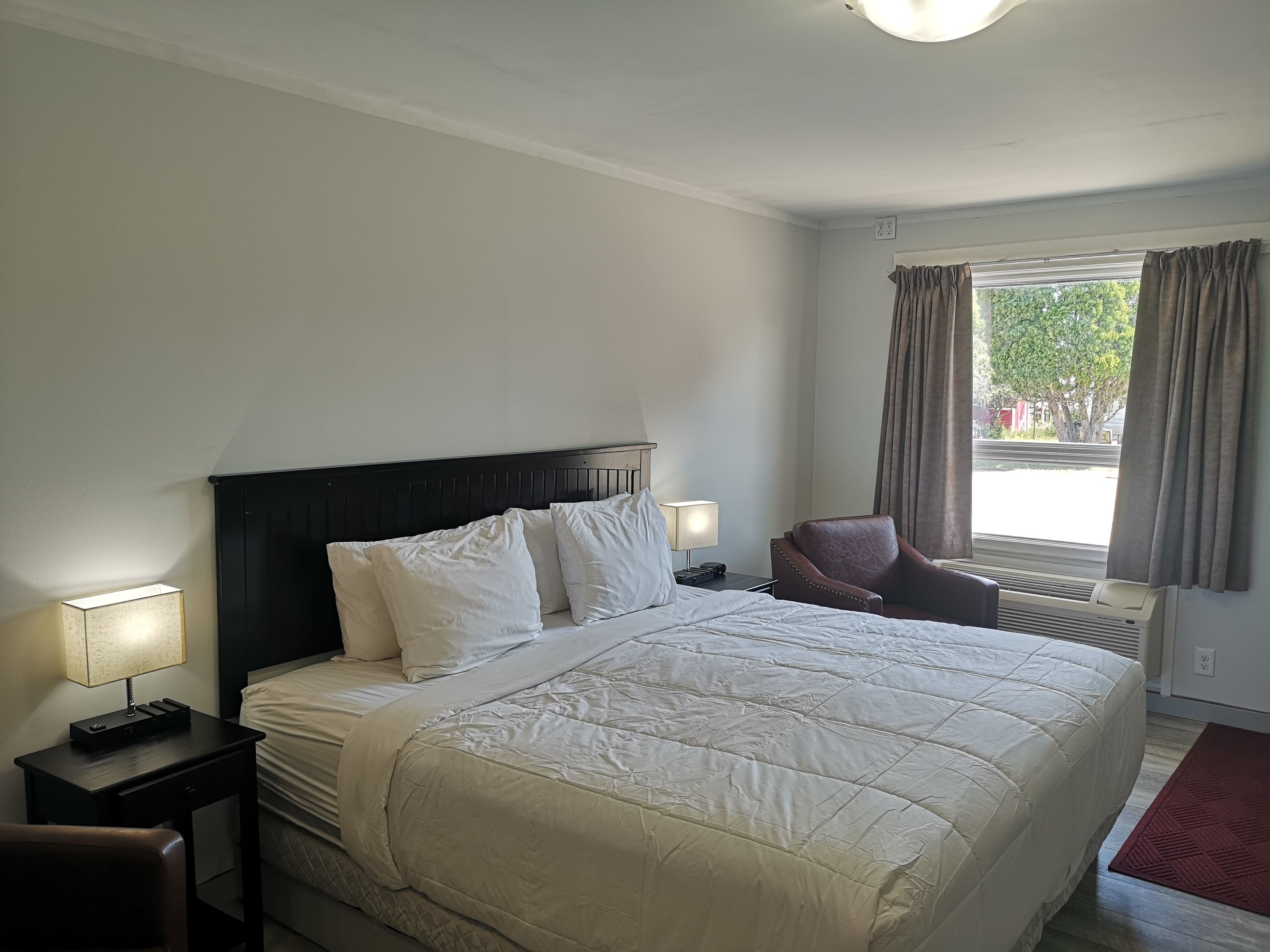Une chambre d’hôtel avec un grand lit, une chaise, un bureau avec une lampe et une fenêtre avec des rideaux.