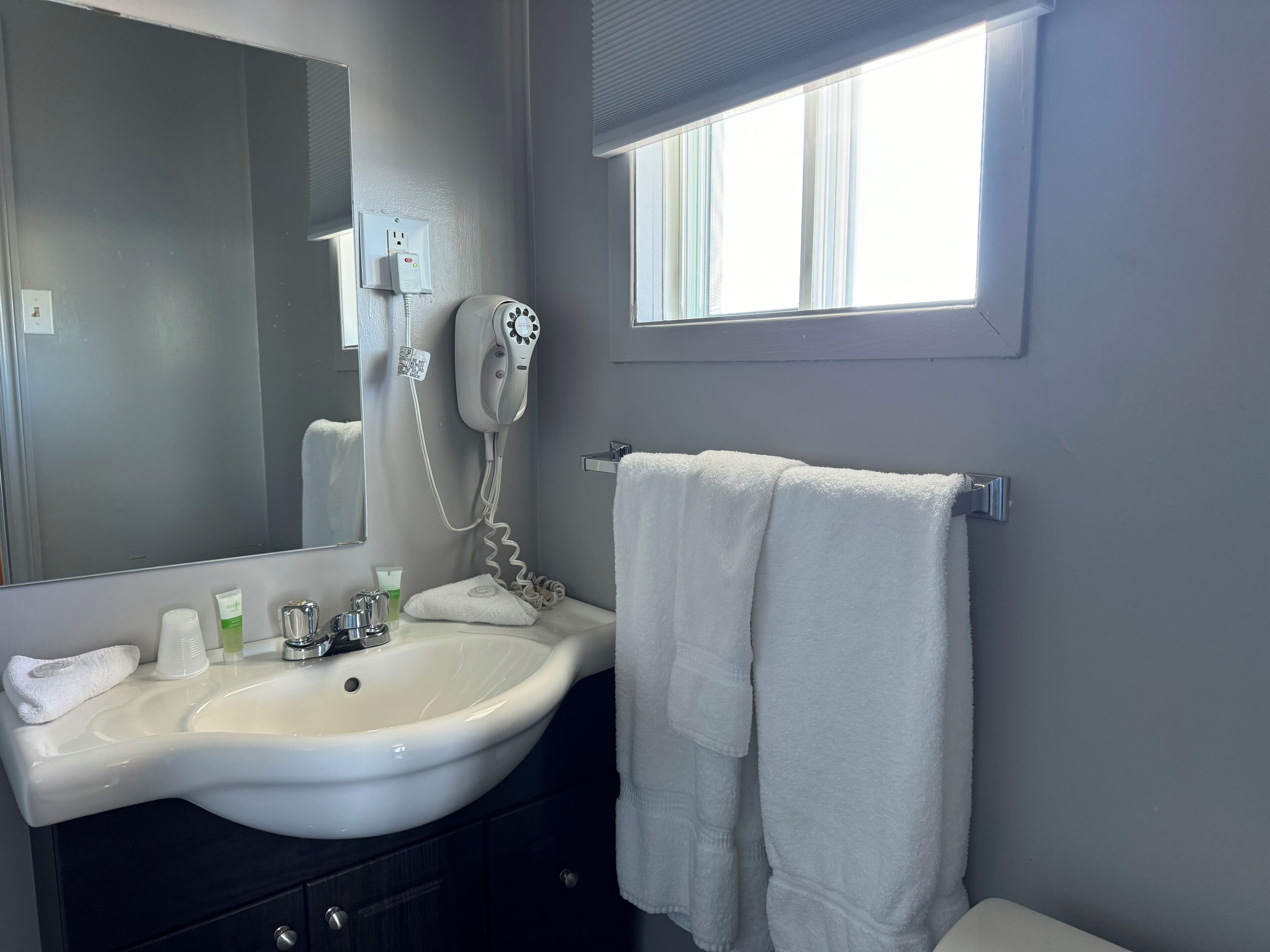 A bathroom with a white sink, a mirror, a hairdryer, a towel, and a window.