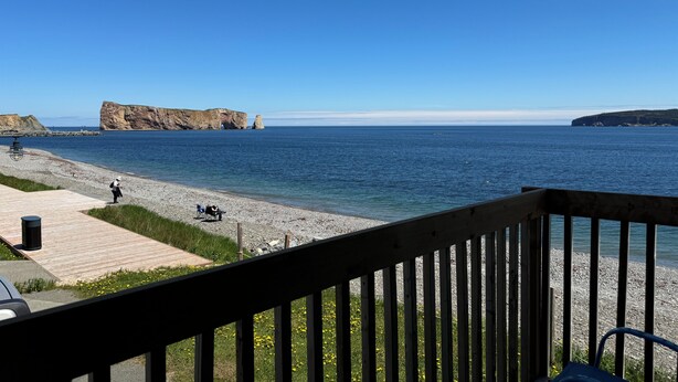 A coastal view from a balcony with a wooden railing, a pebble beach, and a rocky cliff in the distance. - Hotel Motel Fleur De Lys (Perce)