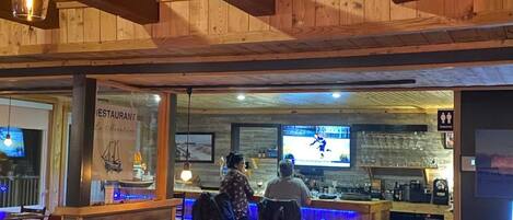 L'intérieur d'un restaurant est agrémenté de poutres en bois, il comprend un bar et un écran de télévision diffusant un événement sportif.