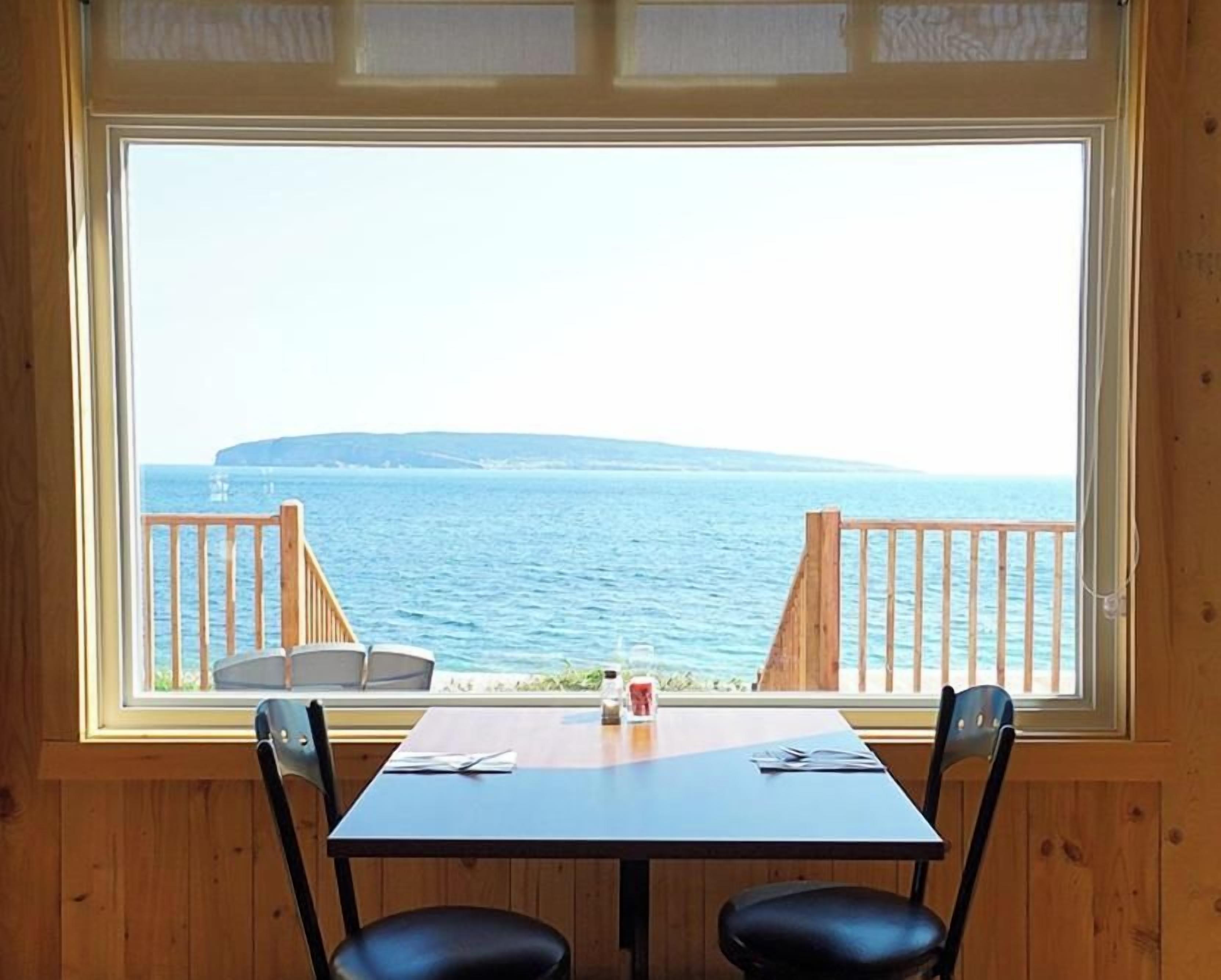 A room with a large window overlooking a body of water and a wooden deck. There is a table with two chairs and a small table with a bottle and a glass.