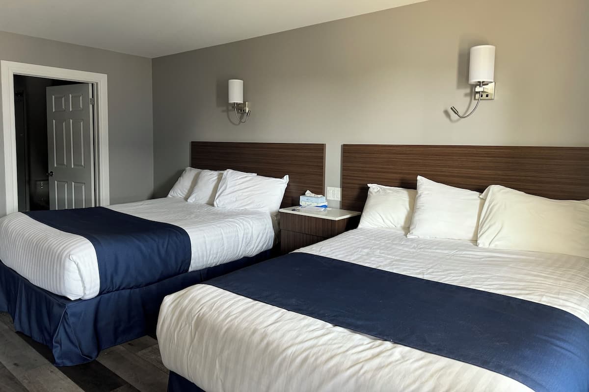A hotel room with two beds, each with a wooden headboard, white bedding, and a blue accent. There are two wall-mounted lamps and a nightstand with a tissue box.
