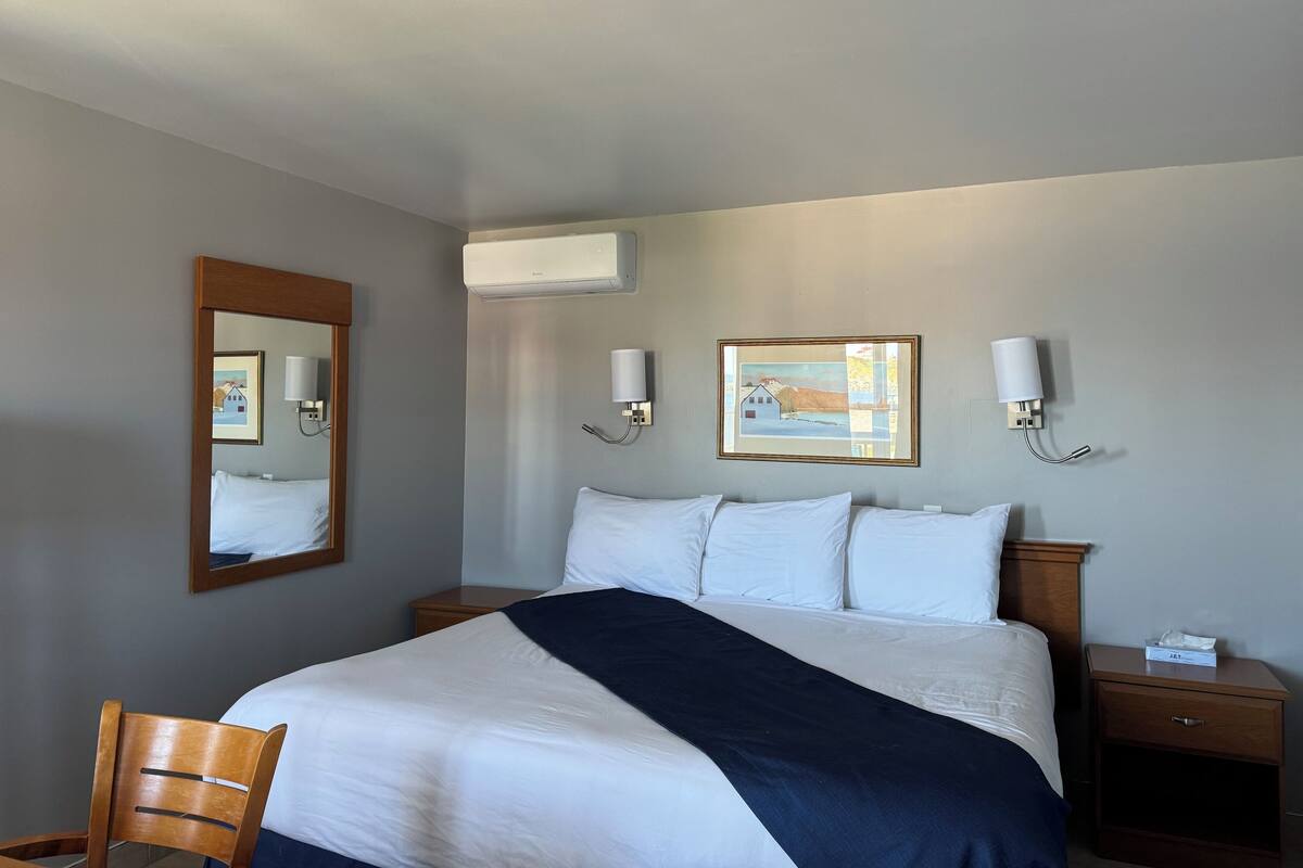 A hotel room with a bed, a chair, a nightstand, an air conditioning unit, and two wall-mounted lamps.