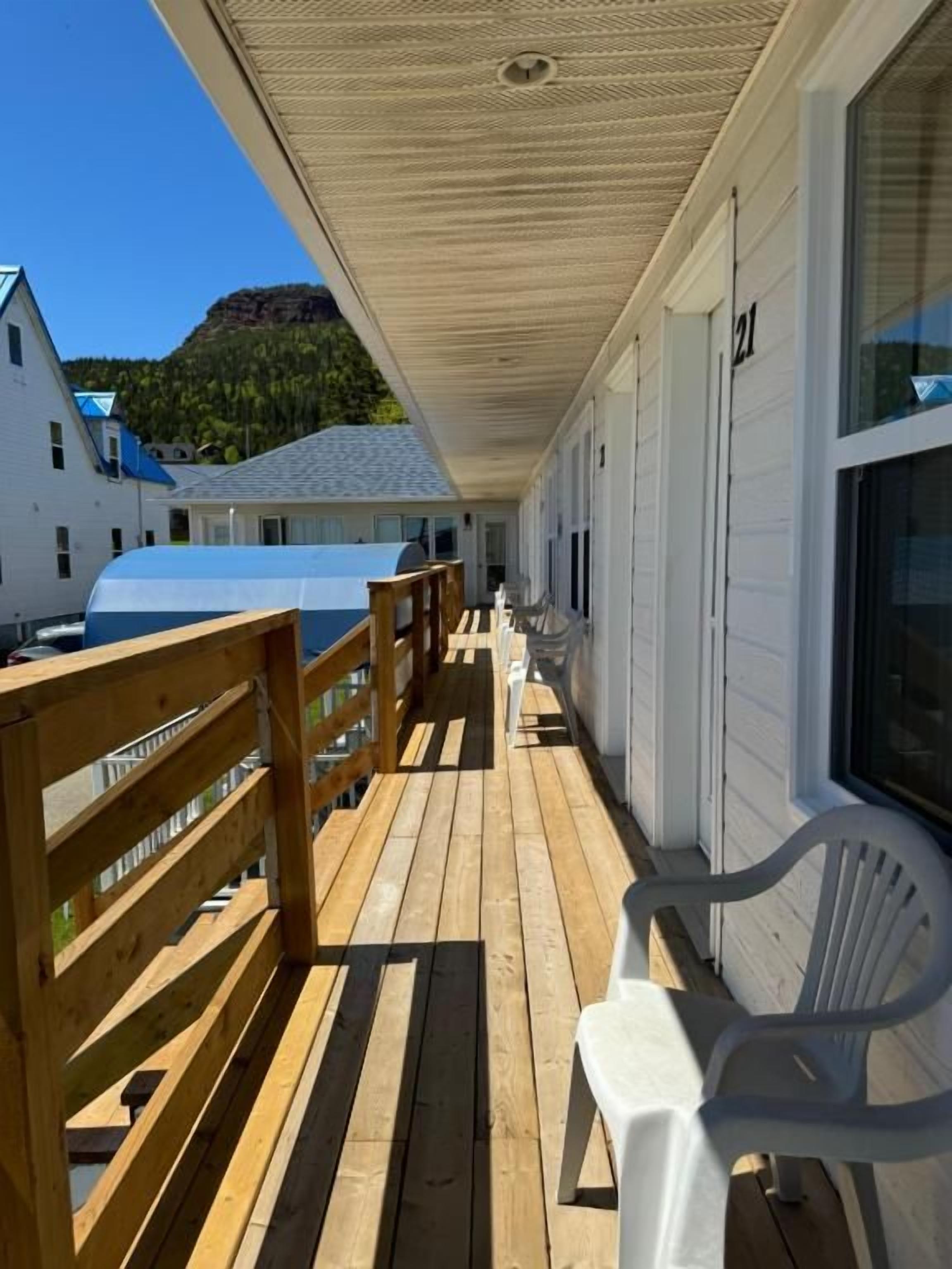 A covered outdoor deck with wooden railings and white plastic chairs, leading to a building with a white exterior and a visible garage door.