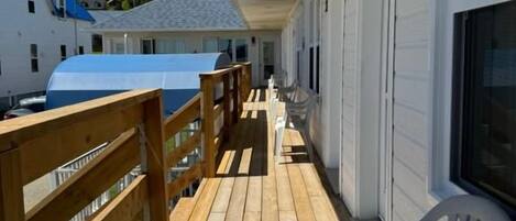 A covered outdoor deck with wooden railings and white plastic chairs, leading to a building with a white exterior and a visible garage door.