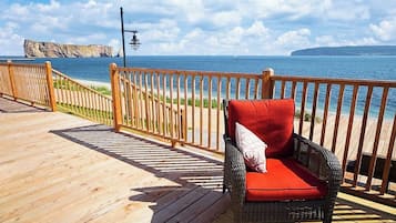 A wooden deck with a wicker chair and a red cushion, overlooking a coastal area with a clear sky.