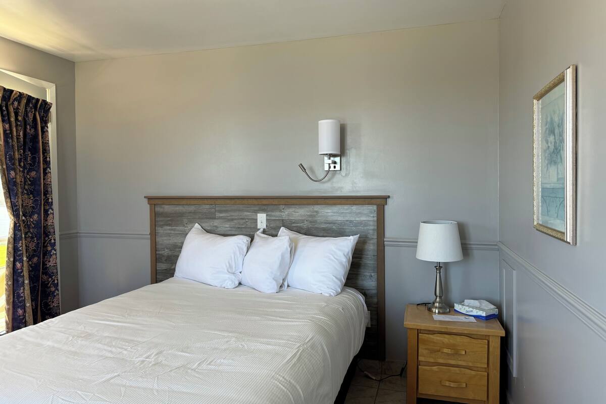 A neatly made bed with white linens, a wooden headboard, a bedside lamp, and a small wooden nightstand with a lamp and a tissue box.