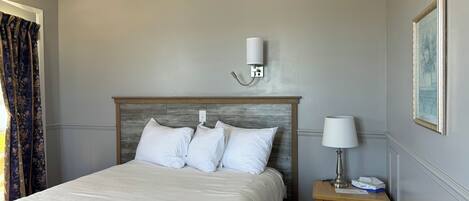 Un letto ben rifatto con lenzuola bianche, una testiera in legno, una lampada da comodino e un piccolo comodino in legno con una lampada e un porta fazzoletti.