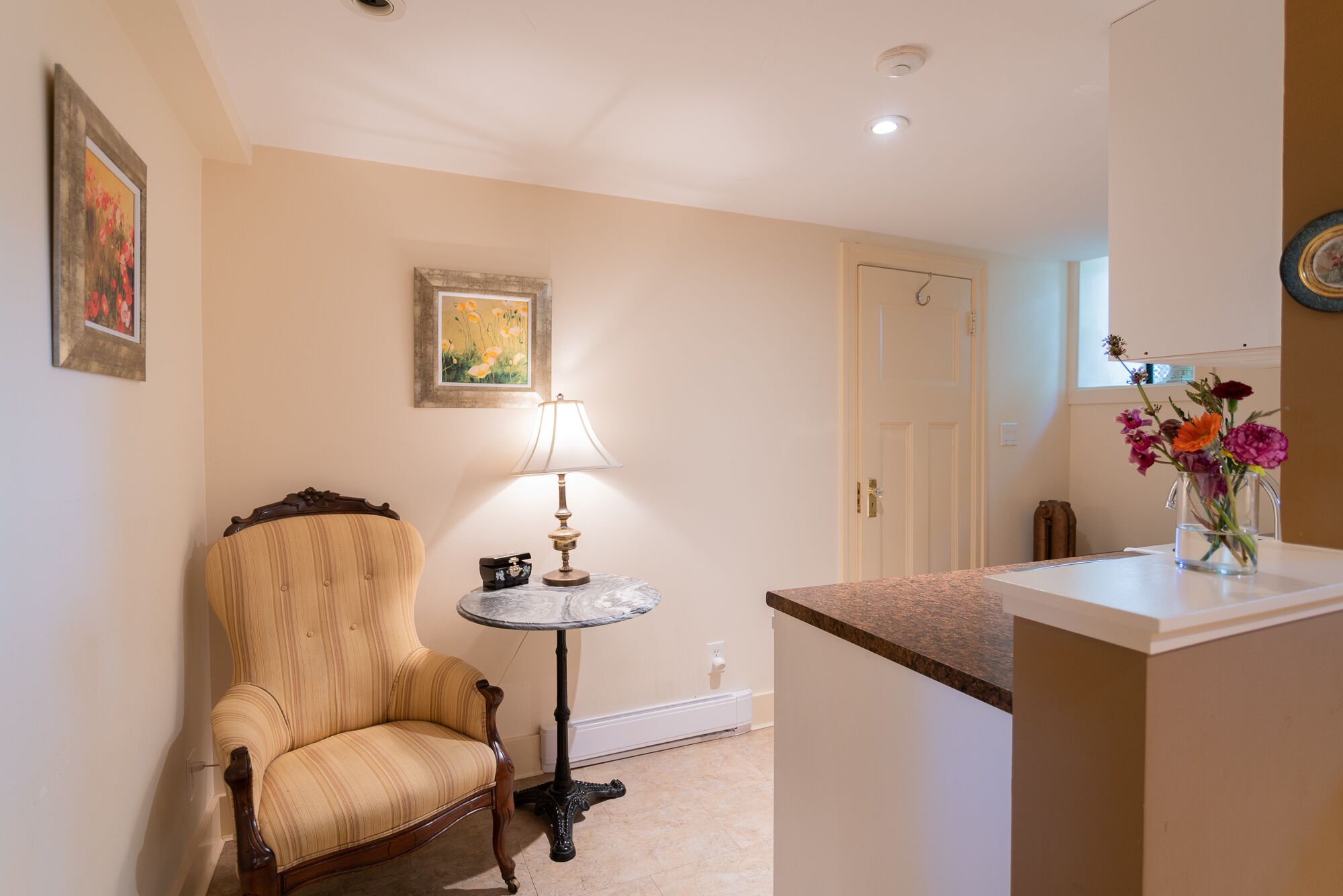 A room with a wooden chair, a small round table with a lamp, and a vase with flowers on a counter.