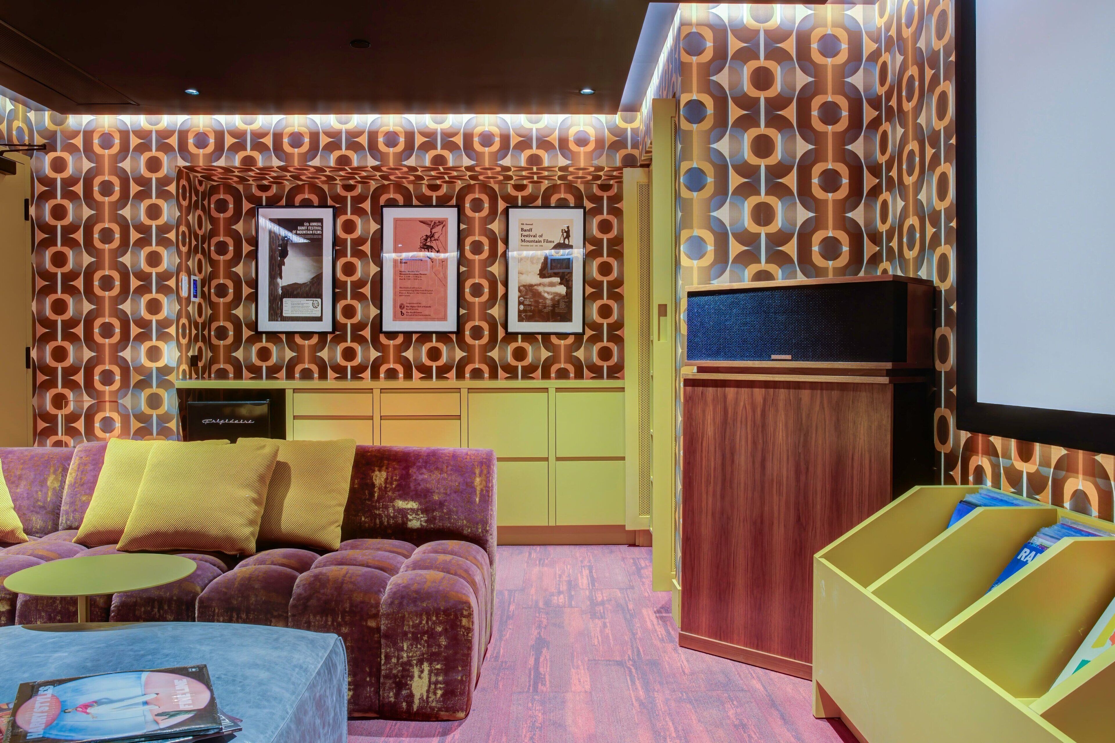 Una habitación con pared estampada, un sofá morado con cojines amarillos, una mesita redonda y un mueble con un televisor.