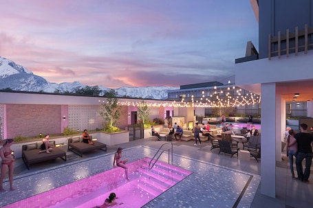 A rooftop pool area with a view of snow-capped mountains, outdoor seating, and ambient lighting.