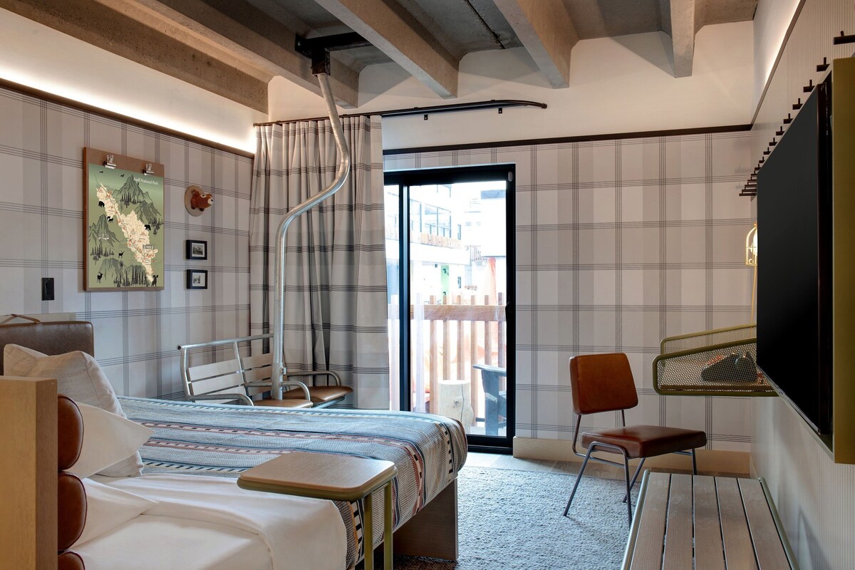A hotel room with a bed, a desk, a chair, a map on the wall, and a view of the outside.
