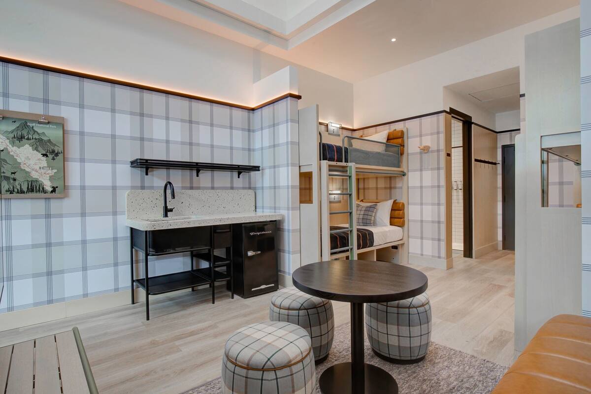 A modern, compact hotel room with a kitchenette, a dining area with ottomans, a bed with a headboard, and a map on the wall.