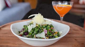 A white bowl of kale salad with pine nuts and shaved cheese on a wooden table.