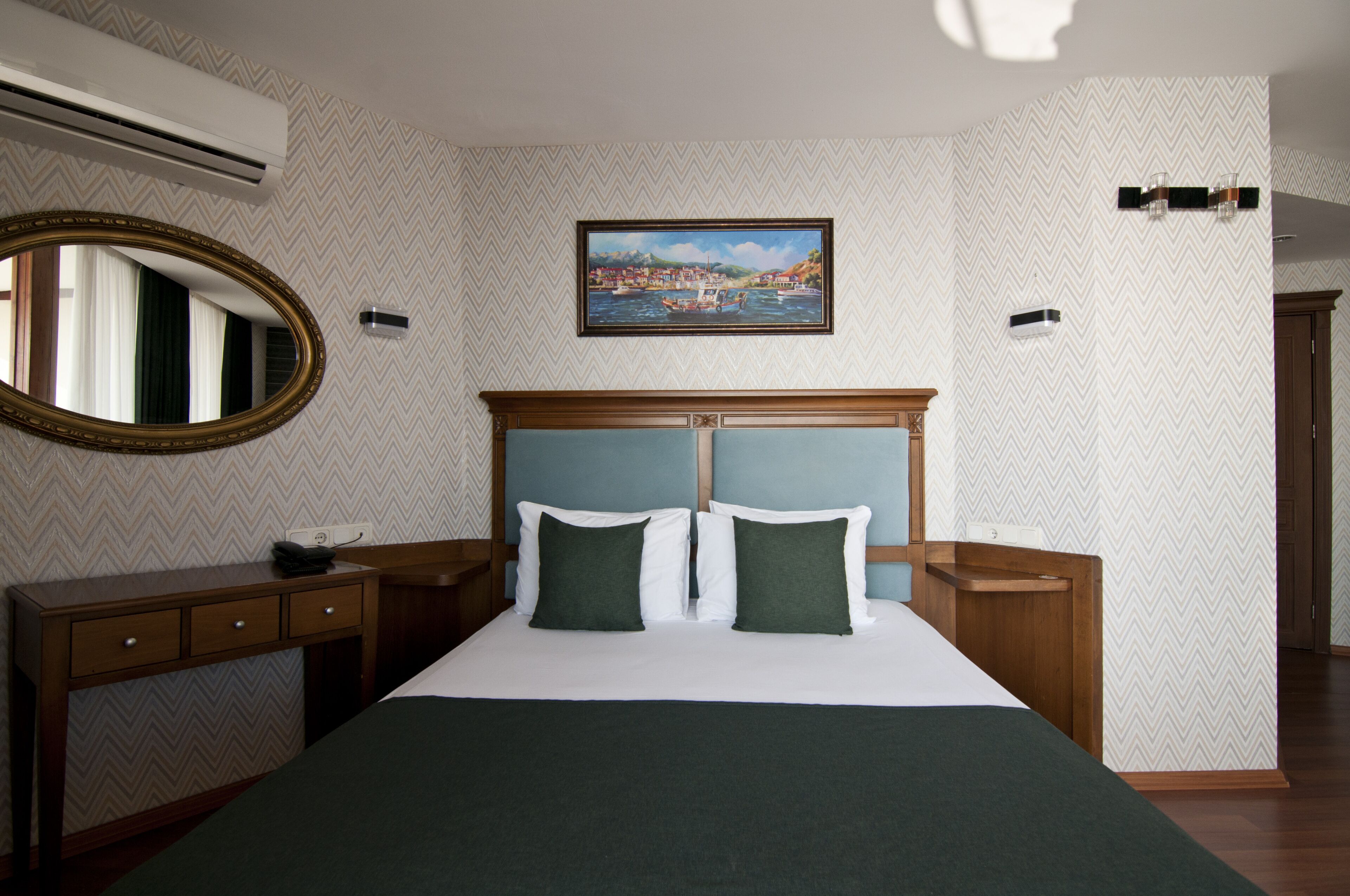 A hotel room with a large bed, a wooden headboard, a bedside table, a mirror, and a painting on the wall.