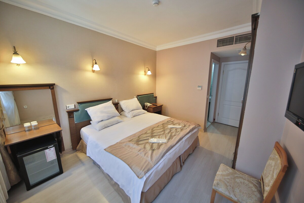 A hotel room with a bed, bedside tables, a chair, a television, and a door leading to another room.