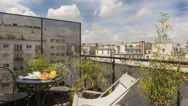 Terraza en la azotea con una mesa preparada para el desayuno, con cruasanes y jugo de naranja, rodeada de sillas y plantas en macetas.