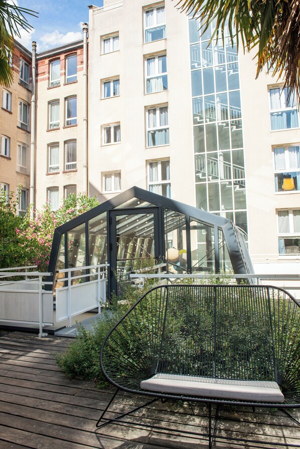 A modern outdoor seating area with a glass enclosure, surrounded by greenery and adjacent to a multi-story building with large windows.