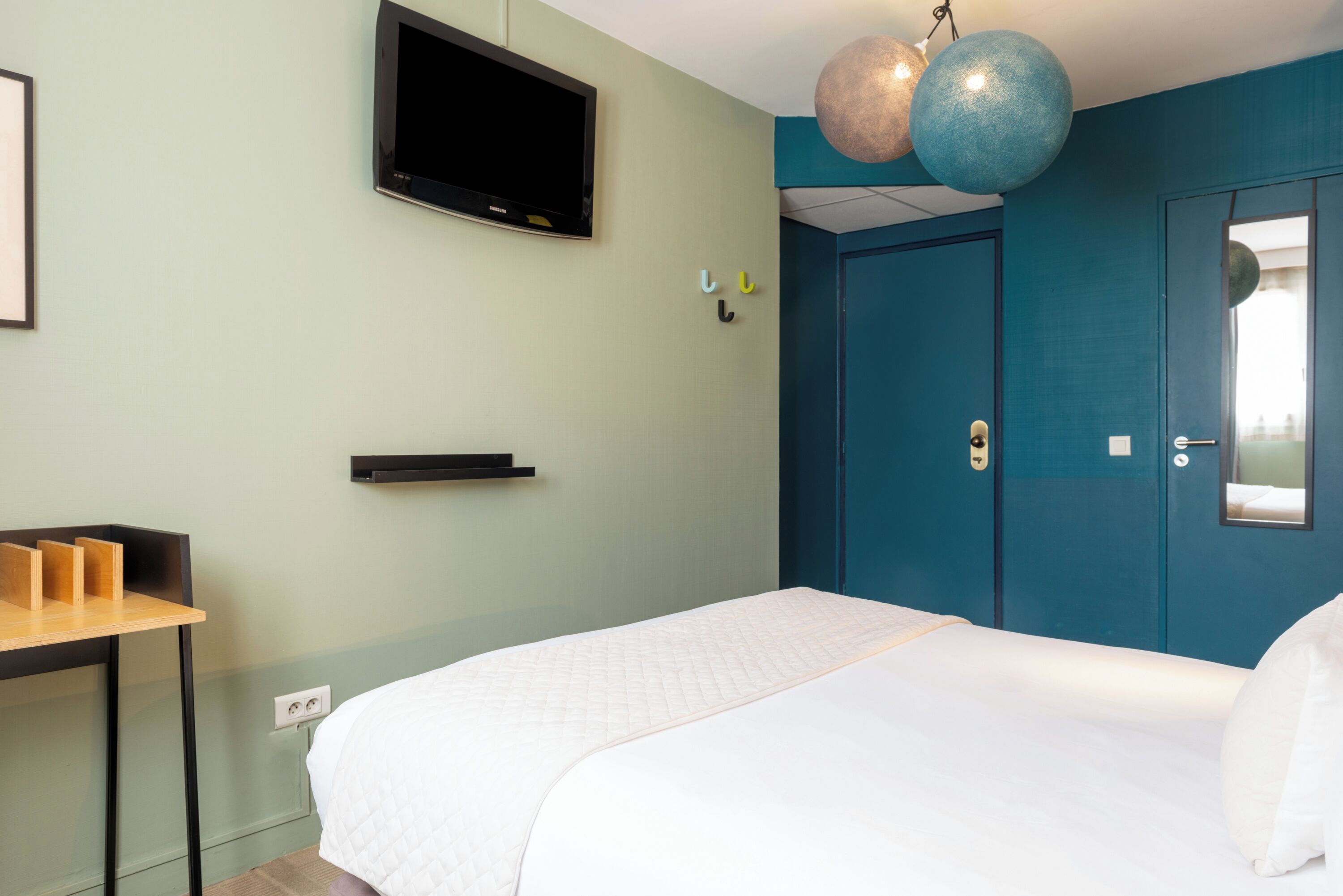 A hotel room with a bed, a TV mounted on the wall, a desk with a chair, and a door with a handle.