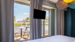 A hotel room with a bed, a TV mounted on the wall, and a balcony with a table and chairs. - Hôtel Bootcamp (Issy-les-Moulineaux)