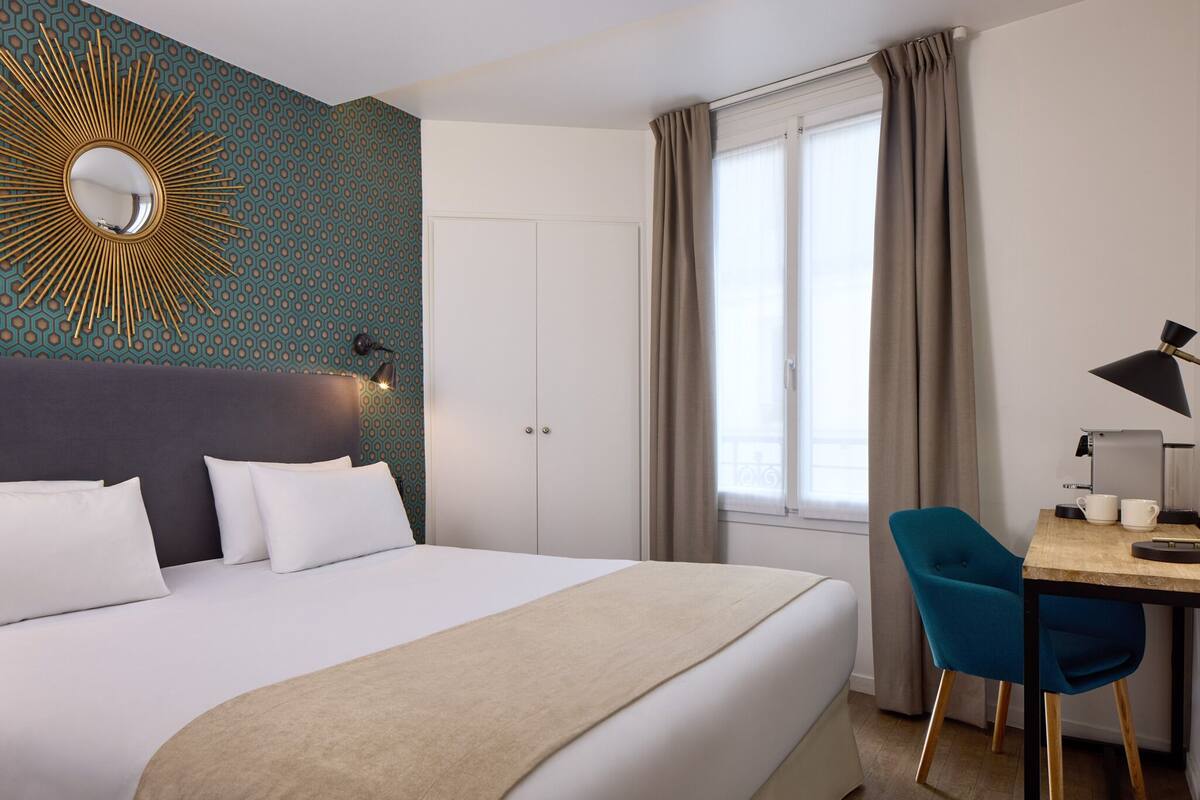 A hotel room with a large bed, a desk with a chair, a window with curtains, and a wall with a decorative mirror and patterned wallpaper.