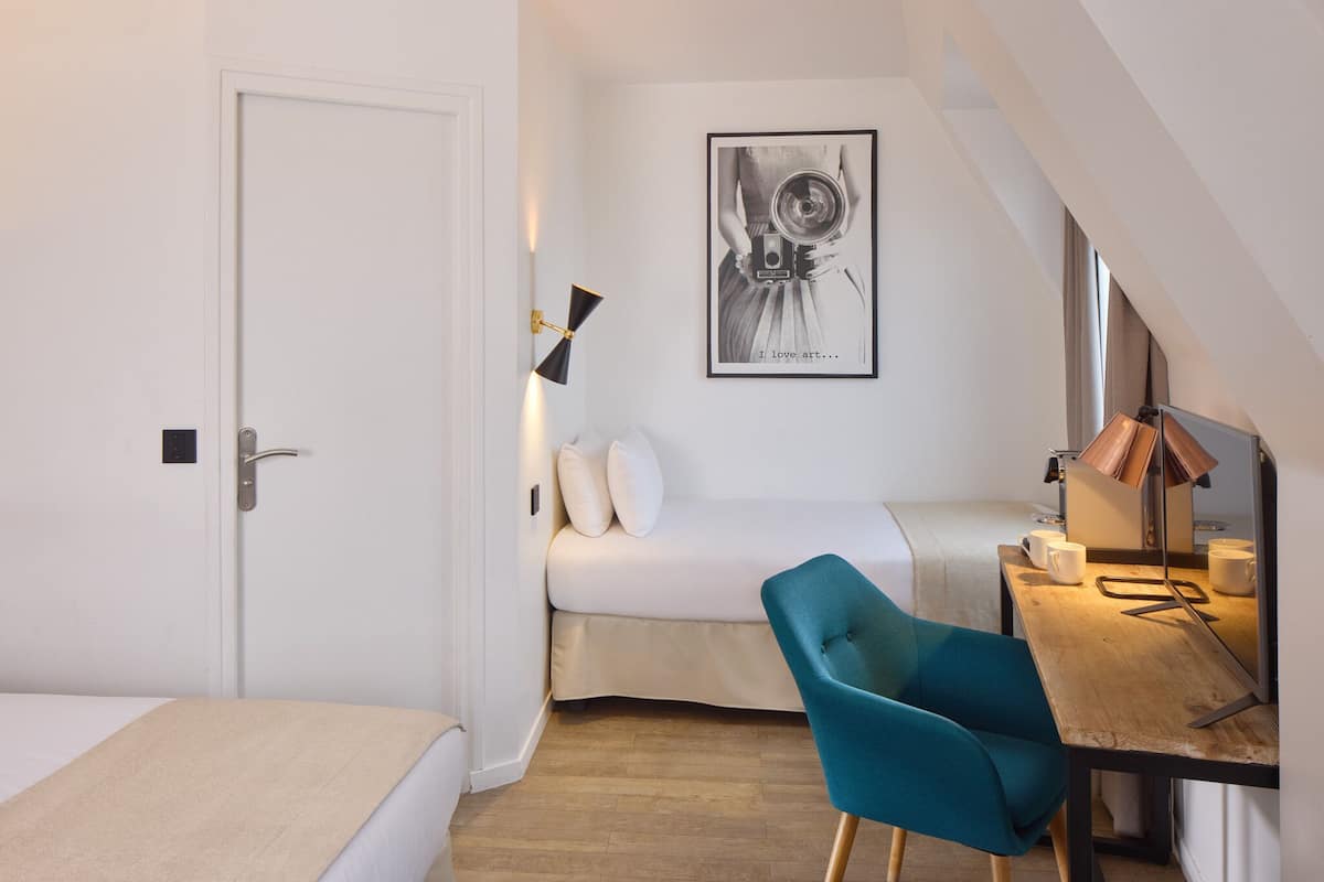 A compact room with a bed, a desk with a chair, a framed artwork on the wall, and a door.