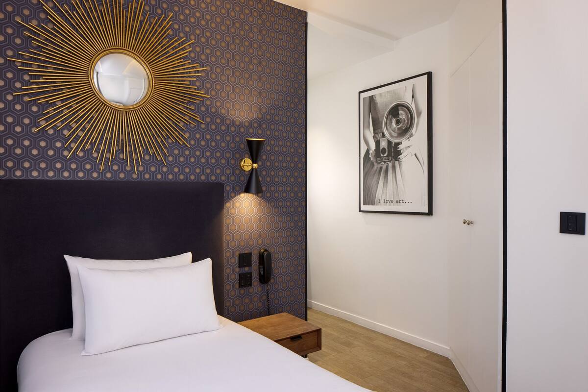 A modern bedroom with a large bed, a wall-mounted mirror, a geometric patterned wall, a framed artwork, and a bedside telephone.