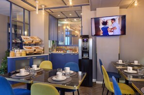 A hotel dining area with tables set for breakfast, a television screen, and a kitchen area visible through a glass partition. - Hotel Ohm by HappyCulture (Paris)