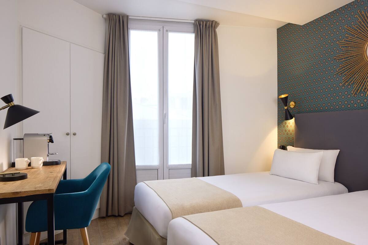 A hotel room with two beds, a desk with a chair, a lamp, and a window with curtains.