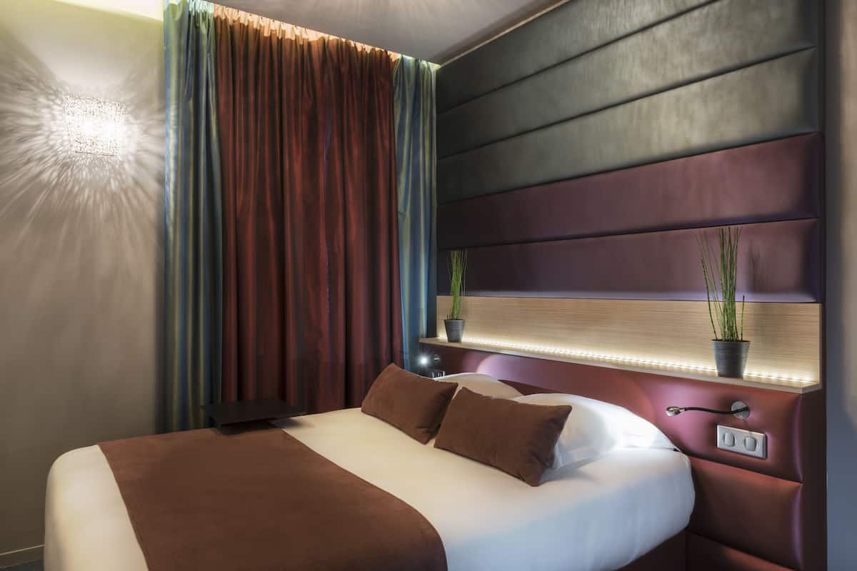 Une chambre d’hôtel moderne avec un grand lit, une tête de lit dotée d’un éclairage intégré, une table de chevet avec une lampe et une plante en pot posée sur l’étagère.