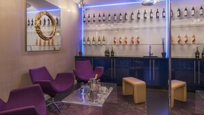 Zona bar moderna con sedie viola, un tavolo in vetro e una bottiglia di champagne, più scaffali pieni di bottiglie di liquore.