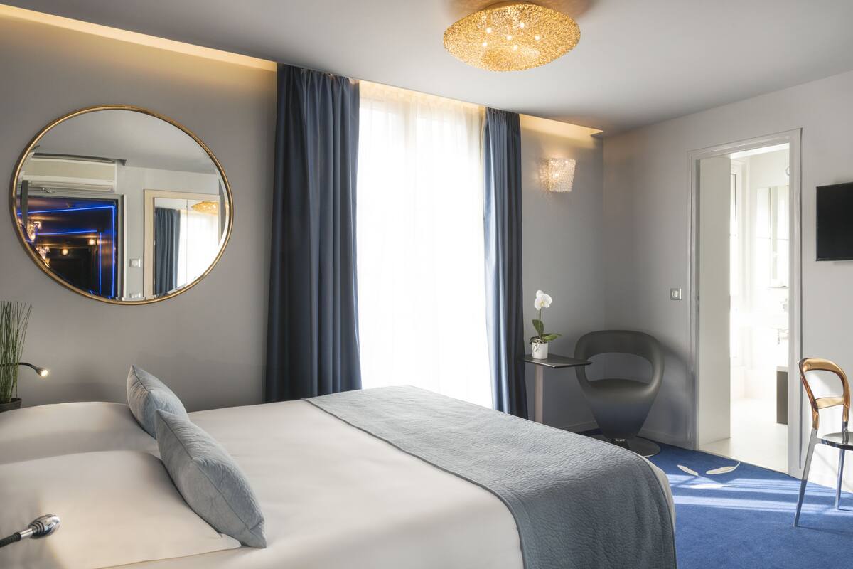 Une chambre d’hôtel moderne avec un grand lit, un miroir rond, un bureau avec une chaise et une vue sur le couloir.