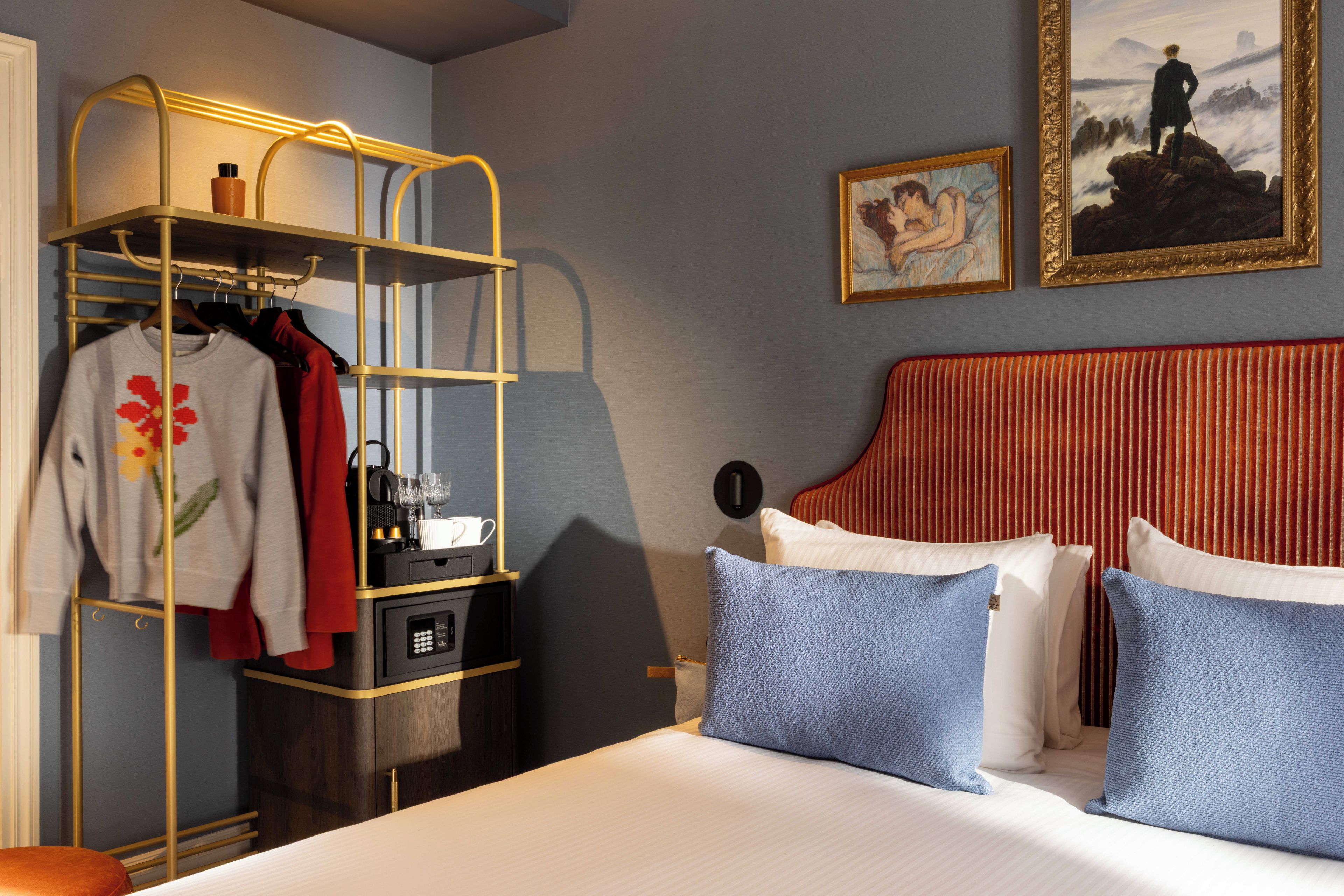 Une chambre d’hôtel avec un lit, une armoire où sont suspendus des vêtements, une machine à café et deux tableaux accrochés au mur.