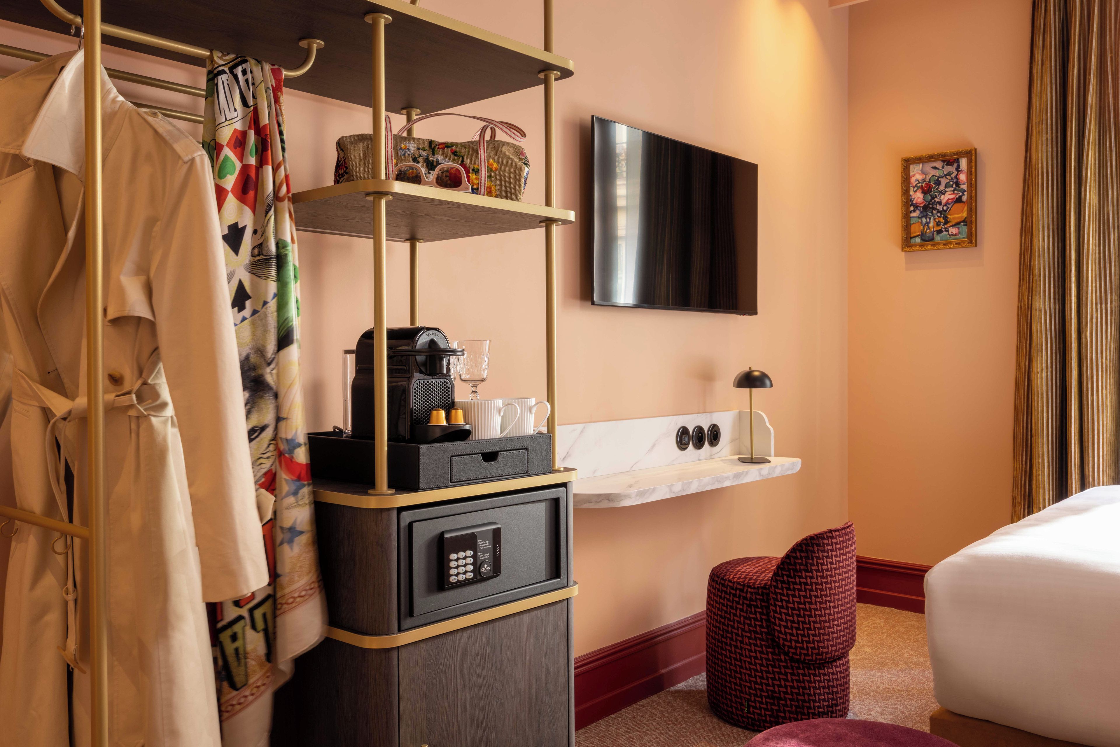 Habitación de hotel con cama, televisor montado en la pared, cafetera, estante con artículos de aseo y una silla roja de fibras.