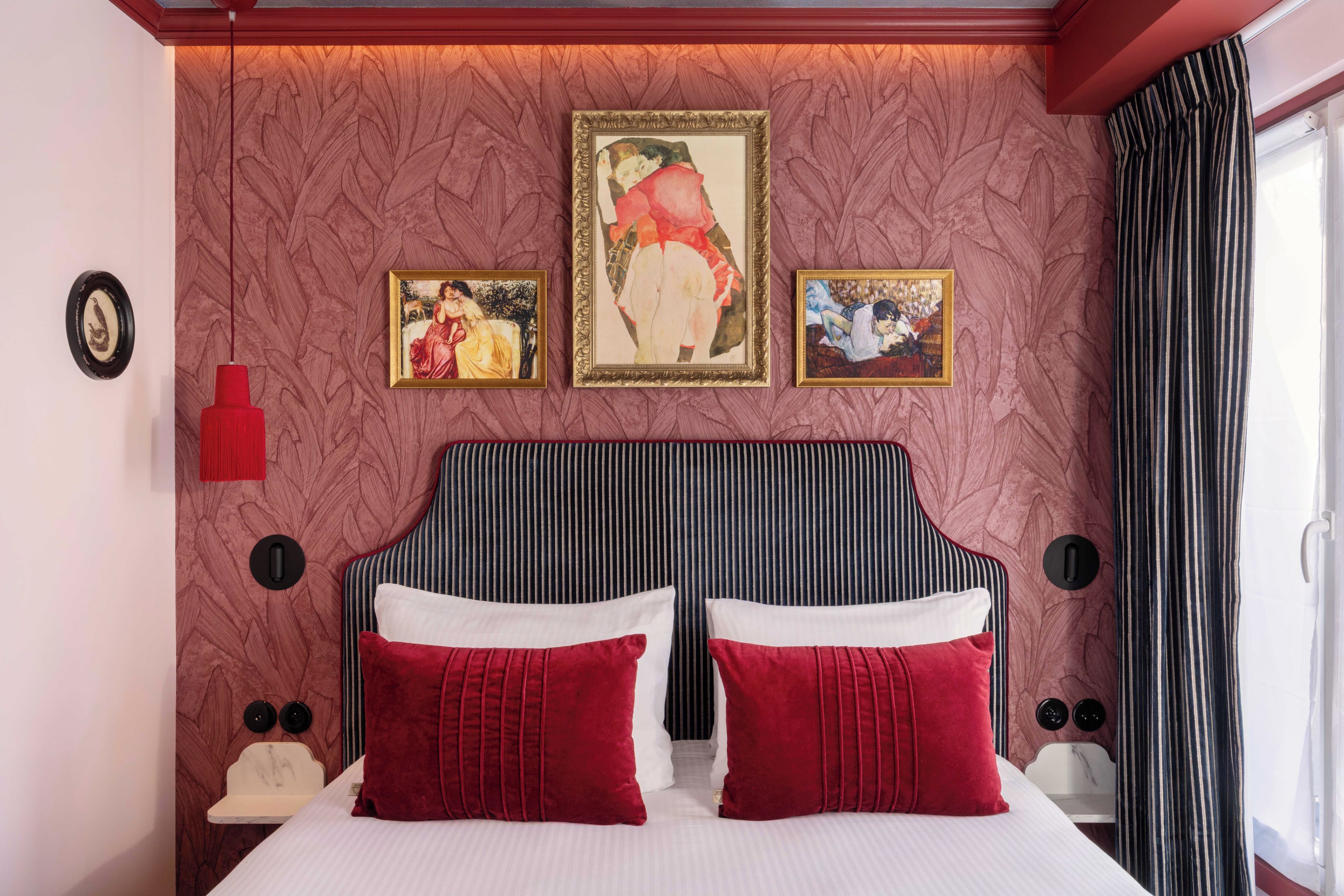 Habitación de hotel con una cama grande, dos almohadas rojas, cabecero con rayas verticales y tres obras de arte enmarcadas en la pared.