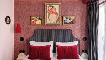 Habitación de hotel con una cama grande, dos almohadas rojas, cabecero con rayas verticales y tres obras de arte enmarcadas en la pared.